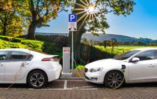 Two plugin electric vehicles charge on a cobblestone street next to open fields on a sunny day