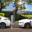 Two plugin electric vehicles charge on a cobblestone street next to open fields on a sunny day