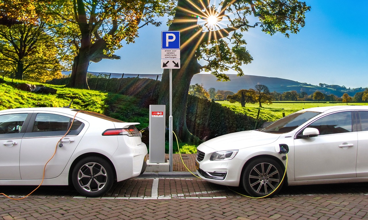 Two plugin electric vehicles charge on a cobblestone street next to open fields on a sunny day